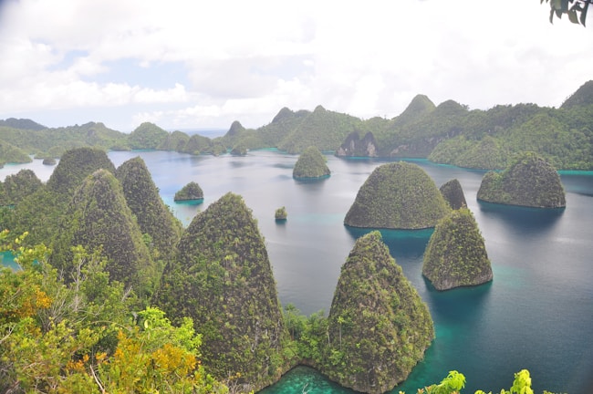 Lush, green cone-shaped islands rise from tropical turquoise waters, surrounded by dense vegetation. Bright skies with scattered clouds form the backdrop. The landscape suggests a serene, remote paradise, with calm waters and a natural, untouched environment.