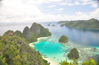 A picturesque landscape featuring lush green islands surrounded by crystal-clear turquoise waters. The islands are covered with dense vegetation, and the water displays various shades of blue and green. A group of people is seen at the edge of one of the islands, enjoying the breathtaking view.