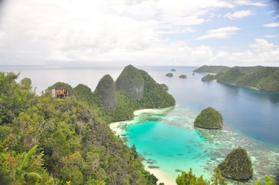A breathtaking view of a tropical archipelago with lush green islands scattered across a turquoise sea. Dense vegetation covers the islands, creating a contrast against the bright blue water. A group of people stands on one of the elevated spots on the island, surrounded by greenery. In the background, the horizon blends into a cloudy sky, adding depth to the scene.