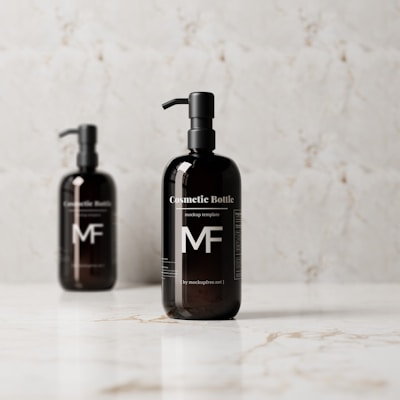 Two dark-colored pump bottles are positioned against a light, marbled background on a reflective surface. The bottles are labeled 'Cosmetic Bottle' and feature a minimalist design with clear, white text and the initials 'MF'.