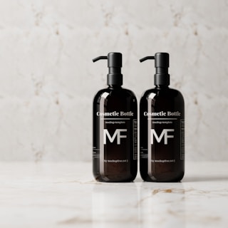 Two identical black cosmetic bottles with pump dispensers are placed side by side on a white marble surface. Each bottle has a minimalist design with prominent 'MF' initials and text indicating they are mockup templates.