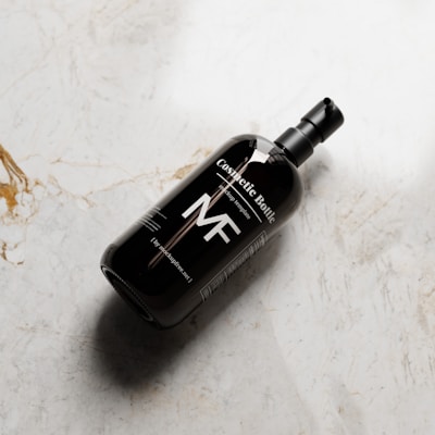 A dark cosmetic bottle with a pump dispenser lies on a textured, light-colored marble surface. The bottle has a minimalist design with bold white text, including the letters 'MF'.