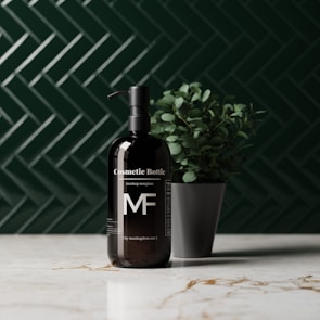 A sleek black cosmetic bottle with a pump sits on a marble countertop. The bottle is labeled with the text 'Cosmetic Bottle' and features the letters 'MF'. It is placed next to a small potted plant with green leaves. The background features a dark green herringbone tile pattern.