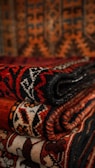 A stack of intricately patterned textiles with geometric designs in rich, warm colors. The patterns feature combinations of reds, blacks, oranges, and whites, showcasing intricate artistic craftsmanship.