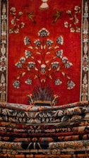 An intricately designed tapestry with a rich, bold red background featuring a central floral motif and decorative birds. The borders are adorned with detailed patterns in contrasting colors. Multiple layers of carpets or textiles, each with their own unique patterns, are stacked in the foreground.