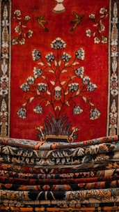An intricately designed tapestry with a rich, bold red background featuring a central floral motif and decorative birds. The borders are adorned with detailed patterns in contrasting colors. Multiple layers of carpets or textiles, each with their own unique patterns, are stacked in the foreground.