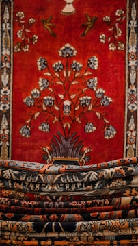 An intricately designed tapestry with a rich, bold red background featuring a central floral motif and decorative birds. The borders are adorned with detailed patterns in contrasting colors. Multiple layers of carpets or textiles, each with their own unique patterns, are stacked in the foreground.
