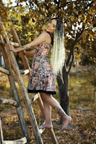 A woman with long, braided hair and floral tattoos is climbing a wooden ladder outdoors. She is wearing a floral dress and barefoot, surrounded by golden autumn leaves and trees.