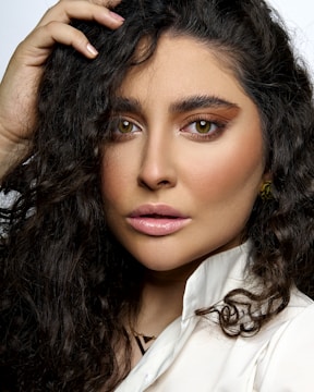 A close-up portrait of a person with long, curly dark hair and expressive brown eyes, framed by well-groomed eyebrows. The person is wearing subtle makeup, including perfectly applied eyeliner and nude lipstick. They are touching their hair with one hand, which has a light pink manicure, while dressed in a white collared shirt. The background is plain, emphasizing the subject's facial features.