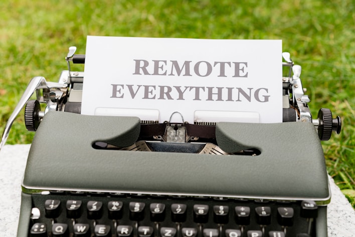 A vintage typewriter is placed on a surface with a background of green grass. A sheet of paper in the typewriter bears the words 'REMOTE EVERYTHING' in bold, uppercase letters. The typewriter has a classic, dark-colored body with visible keys, suggesting a sense of nostalgia and contrast with the modern concept of remote work or connectivity.