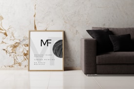 A minimalist interior scene featuring a modern sofa with dark cushions placed against a light marble wall with gold veins. Next to the sofa is a framed artwork or poster with the letters MF and text indicating a mockup for canvas painting or photo. The floor is tiled in a polished light color.