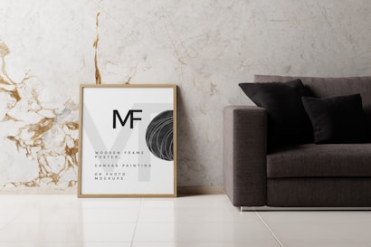 A minimalist interior scene featuring a modern sofa with dark cushions placed against a light marble wall with gold veins. Next to the sofa is a framed artwork or poster with the letters MF and text indicating a mockup for canvas painting or photo. The floor is tiled in a polished light color.