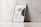 A wooden frame with an abstract poster including the letters 'MF' and design elements like a black circular shape and text detailing uses for wooden frames, canvas paintings, or photo mockups. The frame leans against a white marble wall with subtle vein patterns on a light tiled floor.