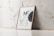 A wooden frame with an abstract poster including the letters 'MF' and design elements like a black circular shape and text detailing uses for wooden frames, canvas paintings, or photo mockups. The frame leans against a white marble wall with subtle vein patterns on a light tiled floor.