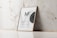 A wooden frame with an abstract poster including the letters 'MF' and design elements like a black circular shape and text detailing uses for wooden frames, canvas paintings, or photo mockups. The frame leans against a white marble wall with subtle vein patterns on a light tiled floor.