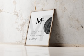 A wooden frame with an abstract poster including the letters 'MF' and design elements like a black circular shape and text detailing uses for wooden frames, canvas paintings, or photo mockups. The frame leans against a white marble wall with subtle vein patterns on a light tiled floor.