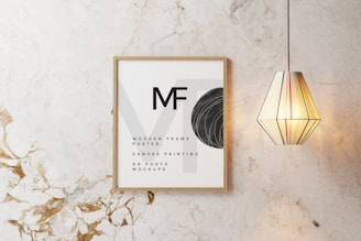 A wooden frame with minimalist text and design elements is displayed on a marble-textured wall. Next to the frame, a geometric pendant light with vertical stripes emits a warm glow.