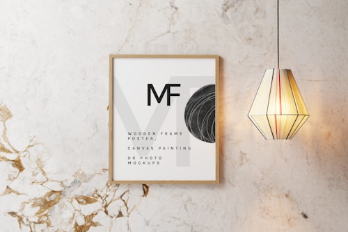 A wooden frame with minimalist text and design elements is displayed on a marble-textured wall. Next to the frame, a geometric pendant light with vertical stripes emits a warm glow.