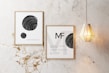 Two framed posters with minimalist black and white designs are displayed on a marble wall. One poster features spheres with swirling patterns, while the other has text and a similar sphere design. A modern, geometric pendant light hangs beside the frames.