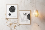 Two framed posters with minimalist black and white designs are displayed on a marble wall. One poster features spheres with swirling patterns, while the other has text and a similar sphere design. A modern, geometric pendant light hangs beside the frames.