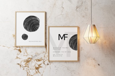 Two framed posters with minimalist black and white designs are displayed on a marble wall. One poster features spheres with swirling patterns, while the other has text and a similar sphere design. A modern, geometric pendant light hangs beside the frames.