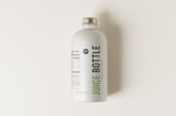 A minimalist juice bottle featuring a white label with nutritional facts and branding. The label includes the words 'Juice Bottle' in bold, with contrasting green and black text. The bottle has a metallic screw cap and is placed on a light, neutral background.