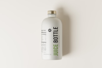 A minimalist juice bottle featuring a white label with nutritional facts and branding. The label includes the words 'Juice Bottle' in bold, with contrasting green and black text. The bottle has a metallic screw cap and is placed on a light, neutral background.