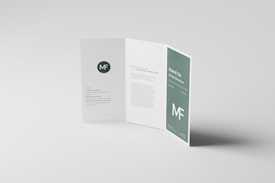 A clean and minimalist tri-fold brochure displayed on a light gray surface. The brochure is partially open, revealing sections with text and graphical elements. A prominent ‘MF’ logo appears, and there are styled text blocks.
