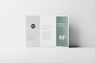 A tri-fold brochure standing open on a minimalistic white surface. The design features a clean, modern layout with a green color theme and white text. The middle panel has a paragraph of text, while the right panel prominently displays the title 'Stand Up Tri-Fold Brochure' and the abbreviation 'MF' in large font size. The left panel contains a circular logo with the initials 'MF' and some smaller text below it.