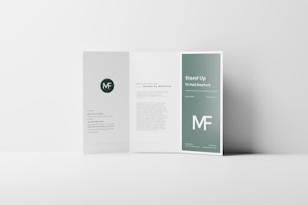 A tri-fold brochure standing open on a minimalistic white surface. The design features a clean, modern layout with a green color theme and white text. The middle panel has a paragraph of text, while the right panel prominently displays the title 'Stand Up Tri-Fold Brochure' and the abbreviation 'MF' in large font size. The left panel contains a circular logo with the initials 'MF' and some smaller text below it.