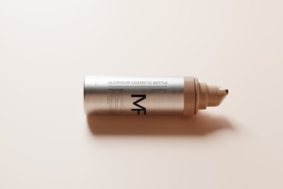 An aluminum cosmetic bottle lying horizontally on a light surface. The bottle has a metallic finish with clear labeling and a nozzle at one end. The prominent text on the bottle reads 'Aluminum Cosmetic Bottle'.