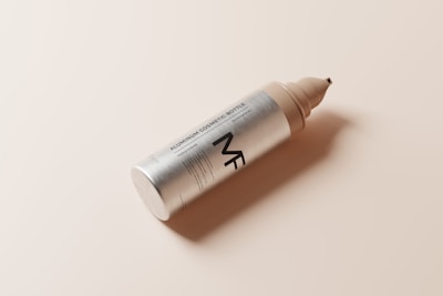 A sleek aluminum cosmetic bottle with a tapered nozzle and minimalist design lies on a smooth, beige surface. The bottle features simple, bold typography, displaying a contemporary aesthetic.
