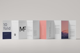 A modern, minimalistic folded brochure with multiple panels displayed in a row. Each panel features a different design, including text elements and an image of a mountain landscape during sunset on one side. The color scheme is predominantly white, with accents of dark blue, black, and soft pink.