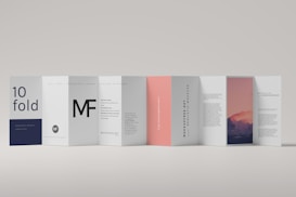 A modern, minimalistic folded brochure with multiple panels displayed in a row. Each panel features a different design, including text elements and an image of a mountain landscape during sunset on one side. The color scheme is predominantly white, with accents of dark blue, black, and soft pink.