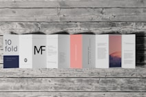 An accordion-style brochure lays open on a wooden surface. The brochure features various panels with text in different fonts and a photograph of a mountain landscape under a pink and purple sky. The text includes large letters, some branding, and mock text representing informational content. One panel is notably a subtle pink color.
