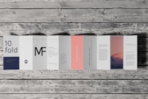 An accordion-style brochure lays open on a wooden surface. The brochure features various panels with text in different fonts and a photograph of a mountain landscape under a pink and purple sky. The text includes large letters, some branding, and mock text representing informational content. One panel is notably a subtle pink color.