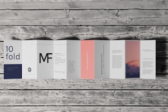 An accordion-style brochure lays open on a wooden surface. The brochure features various panels with text in different fonts and a photograph of a mountain landscape under a pink and purple sky. The text includes large letters, some branding, and mock text representing informational content. One panel is notably a subtle pink color.