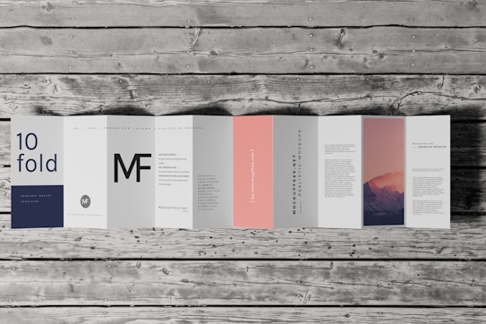 An accordion-style brochure lays open on a wooden surface. The brochure features various panels with text in different fonts and a photograph of a mountain landscape under a pink and purple sky. The text includes large letters, some branding, and mock text representing informational content. One panel is notably a subtle pink color.