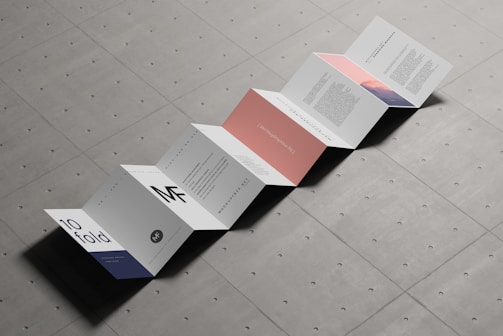 An accordion-style brochure is laid out on a textured surface. The brochure consists of multiple panels with a minimalist design, featuring text and some graphical elements in muted colors. Each panel displays different content, with some having bold letters and others displaying paragraphs of text.
