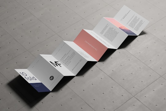 An accordion-style brochure is laid out on a textured surface. The brochure consists of multiple panels with a minimalist design, featuring text and some graphical elements in muted colors. Each panel displays different content, with some having bold letters and others displaying paragraphs of text.