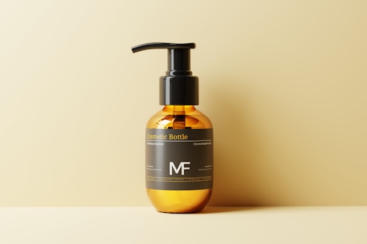 A sleek amber-colored cosmetic bottle with a black pump dispenser is placed against a soft beige background. The label on the bottle features minimalist typography with the initials 'MF' prominently displayed.