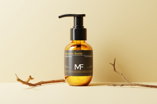 A cosmetic bottle with a pump top is placed on a cream-colored surface and background. The bottle is amber-colored with a black label featuring text and a monogram. Two small, dry branches are laid beside the bottle, adding to the minimalist aesthetic.