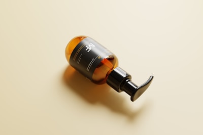 A sleek amber bottle with a black pump nozzle, resting on its side against a light beige background. The bottle has a minimalist black label with white text, reflecting a modern and clean design.
