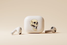 A pair of wireless earbuds and their charging case are displayed. The charging case features a prominent design of a detailed skull illustration on its front. The earbuds are positioned on either side of the case, all set against a soft, beige background.