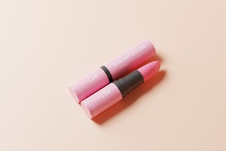 A pink lipstick in an open tube lies on a soft, light beige surface. The text on the tube is visible, and the lipstick is partially extended, ready for application. The composition is minimalistic and elegant.