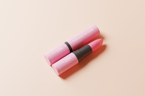A pink lipstick in an open tube lies on a soft, light beige surface. The text on the tube is visible, and the lipstick is partially extended, ready for application. The composition is minimalistic and elegant.