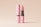A pink tube of lipstick stands next to its matching lid with branding text. The lipstick is partially extended, revealing its vibrant pink color. The background is a soft, neutral beige, creating a minimalist and clean aesthetic.