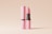 A pink tube of lipstick stands next to its matching lid with branding text. The lipstick is partially extended, revealing its vibrant pink color. The background is a soft, neutral beige, creating a minimalist and clean aesthetic.