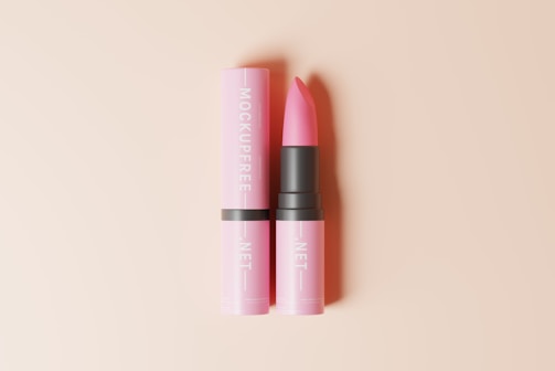 A pink lipstick is positioned against a soft beige background. The lipstick is housed in a sleek, cylindrical case with a matching pink cover that is placed beside it. The words 'Mockupfree.net' are printed vertically on the cover in a contrasting white font.