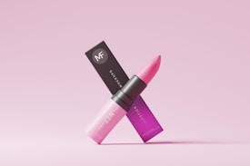 A pink lipstick is positioned in an elegant manner along with its packaging. The lipstick tube is sleek and modern, with a gradient of pink shades transitioning from light at the bottom to a darker hue at the top. The packaging displays the brand logo along with text in a matching pink color scheme. The overall aesthetic is minimalist and chic, set against a light pink background.